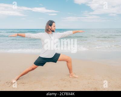 Sportive bearded male training on tranquil seashore and doing yoga asana against blue sea and sky Stock Photo