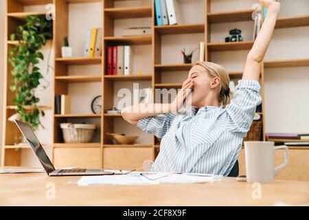Image of sleepy blonde woman yawning while working with papers and laptop at table in office Stock Photo