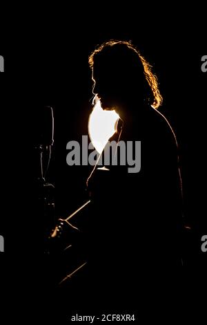 Male guitarist with long hair sitting in dark studio near microphone and glowing lamp and playing guitar Stock Photo