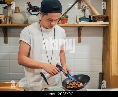 Concentrated young man frying ingredients for Japanese dish called ramen in Asian restaurant Stock Photo