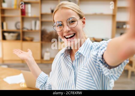Image of cheerful attractive woman in eyeglasses holding copyspace while taking selfie photo at office Stock Photo