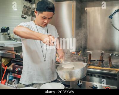 Young man in black cap pouring hot broth from saucepan for Japanese dish called ramen in Asian cafe Stock Photo