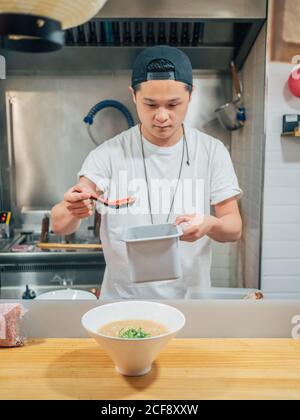 Asian male adding chopped green onions in bowl with fresh cooked Japanese dish on wooden table Stock Photo