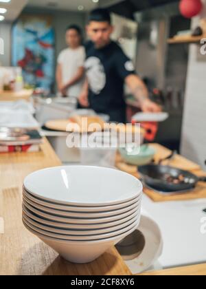 Stack of white bowls prepared for serving ramen on wooden counter and blurred male cooks in kitchen Stock Photo