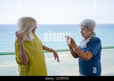 Cheerful mature lady in stylish outfit smiling and taking picture of old friend while standing on terrace near waving sea on resort Stock Photo