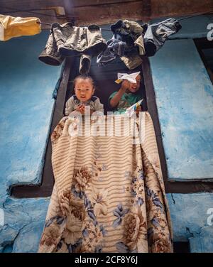 NEPAL, PASHUPATINATH TEMPLE - AUGUST 4, 2014: From below Indian children with paper planes leaning out from window of old building near cloths hanging on twist Stock Photo