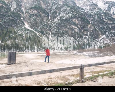 Side view of man in warm red jacket taking photo of amazing majestic landscape of tree and rocky mountains Stock Photo
