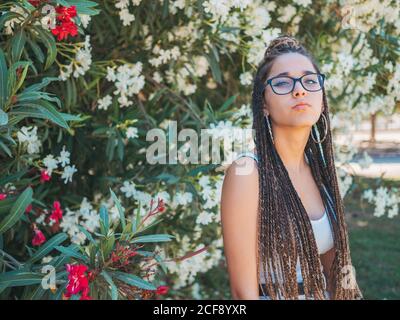 Beautiful young Woman in glasses summer clothes and with dreadlocks standing near blooming trees and looking at camera Stock Photo