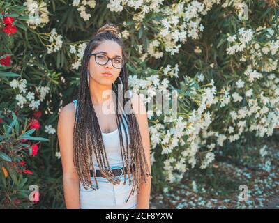 Beautiful young Woman in glasses summer clothes and with dreadlocks standing near blooming trees and looking at camera Stock Photo