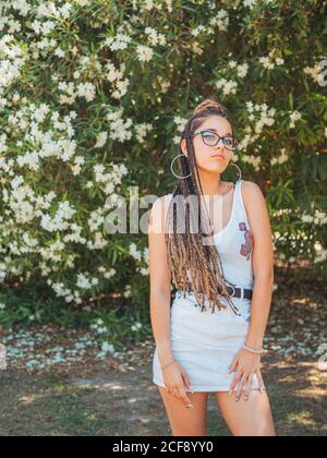 Beautiful young Woman in glasses summer clothes and with dreadlocks standing near blooming trees and looking at camera Stock Photo