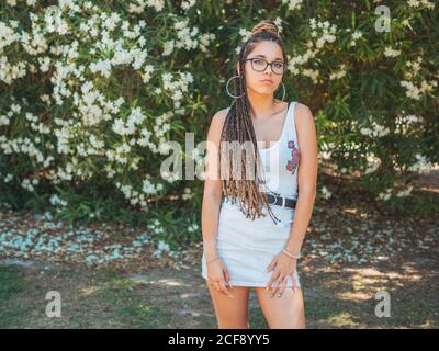 Beautiful young Woman in glasses summer clothes and with dreadlocks standing near blooming trees and looking at camera Stock Photo