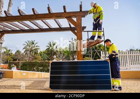 Group of workers in uniform and helmets installing photovoltaic panels on roof of wooden construction near house Stock Photo
