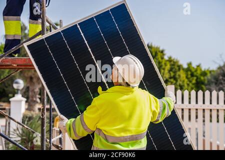 Back view of mature experienced male technician in uniform and helmet standing with solar panel near yellow building while working on installation of renewable energy system Stock Photo