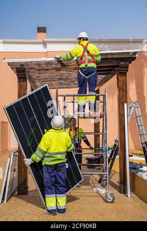 Group of workers in uniform and helmets installing photovoltaic panels on roof of wooden construction near house Stock Photo