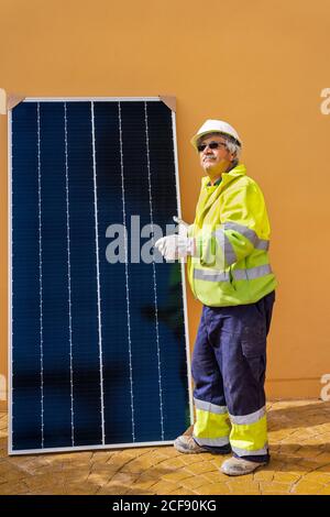 Side view of mature experienced male technician in uniform and helmet standing with solar panel near yellow building while working on installation of renewable energy system Stock Photo