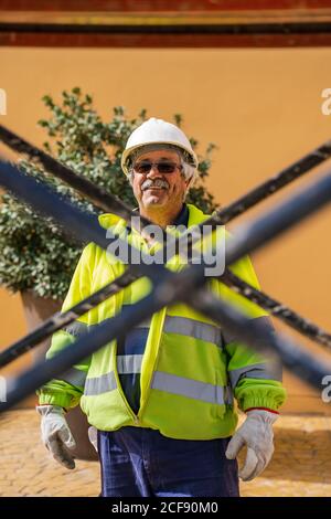 Positive skilled gray haired workman in uniform and protective helmet looking at camera while standing behind metal construction against building wall in sunny day Stock Photo