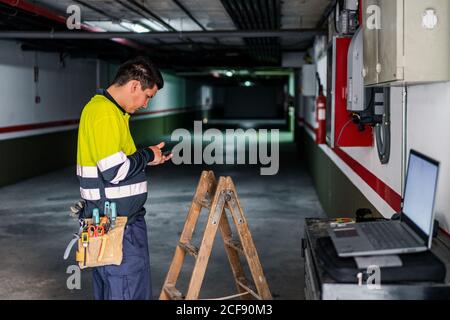 Side view of skilled man engineer in uniform using mobile phone while examining electrical equipment in modern building Stock Photo