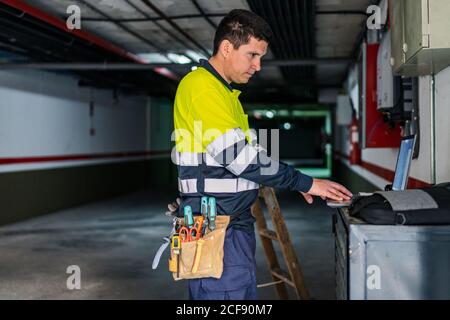 Side view of skilled man engineer in uniform using gadgets while examining electrical equipment in modern building Stock Photo