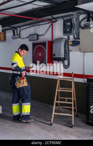 Side view of skilled man engineer in uniform using mobile phone while examining electrical equipment in modern building Stock Photo