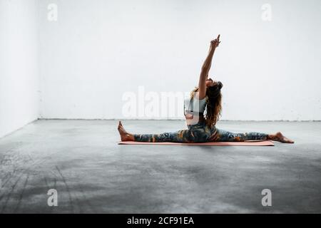 Side view of young flexible female in sportswear doing front split with raised arms while sitting on floor Stock Photo