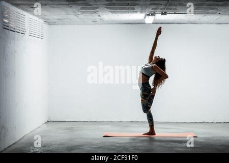 Side view of calm female wearing sports leggings and bra balancing on leg in Urdhva Hastasana and looking up Stock Photo