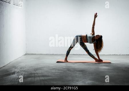 Side view of calm female wearing sports leggings and bra standing on mat in extended triangle pose looking up Stock Photo