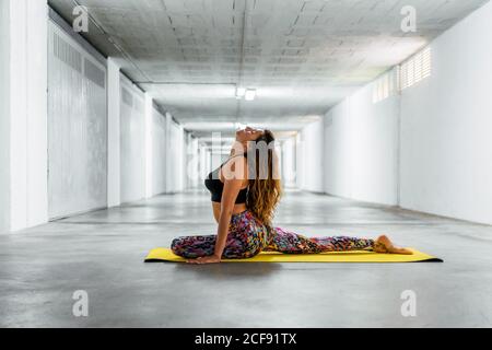 Side view of adult woman in pigeon pose with closed eyes while practicing yoga in light underground hall