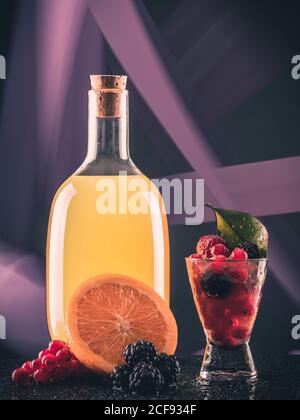 Glass bottle with orange beverage near shot with alcohol and berries on board on abstract background Stock Photo