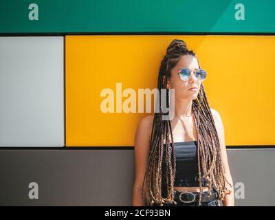 Pretty stylish teenage girl with hands in pocket and unique dreadlocks looking away on colorful background Stock Photo