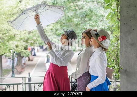 Cheerful Woman in retro dress smiling and opening lace umbrella while standing on terrace in garden near female friends Stock Photo