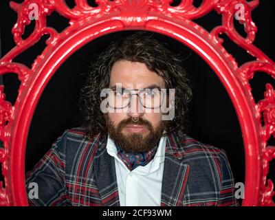Serious impressive well dressed man with curly hair and beard posing in red patterned frame on black background Stock Photo