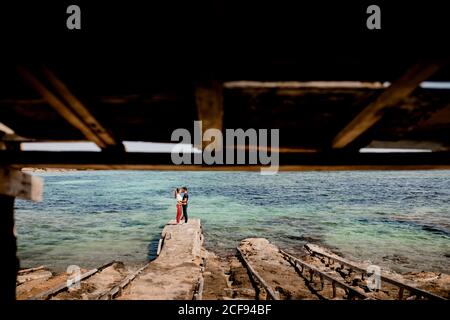 Side view of tender couple at distance embracing on coast with turquoise ocean water in summer time Stock Photo