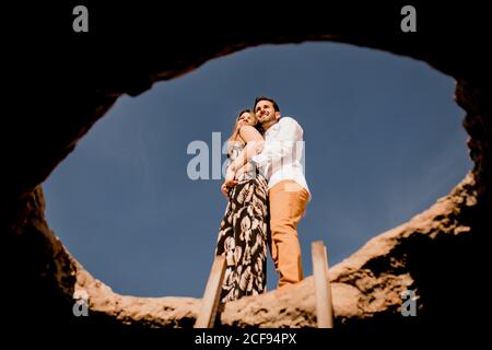 From below of loving young man and Woman in dress hugging on stone well against clear blue sky Stock Photo