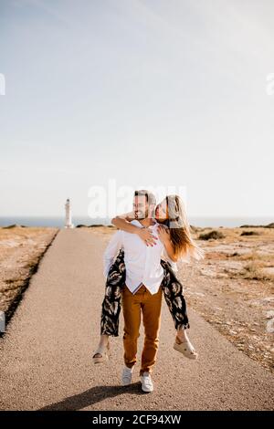 Loving man giving piggyback tide to woman while walking on road from lighthouse and sea in summer and looking away