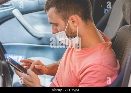 A young man, wearing a cloth face mask, checks the mobile phone inside the car, while he waits for someone before drive, in Spain. Stock Photo