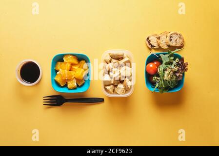 Top view of food containers with bread and tomatoes, arugula and pieces of orange, soy sauce and chicken fillet with black plastic fork on yellow background Stock Photo
