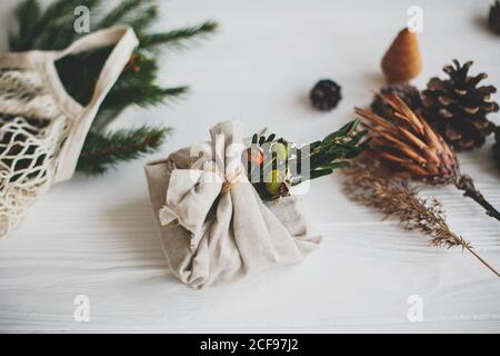 Stylish christmas gift wrapped in linen fabric, decorated with natural green branch on white rustic table background with pine cones and herbs. Zero w Stock Photo