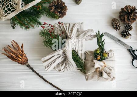 Zero waste stylish christmas gift. Gift wrapped in linen fabric with natural green branch on white rustic table background with fir and scissors. Plas Stock Photo