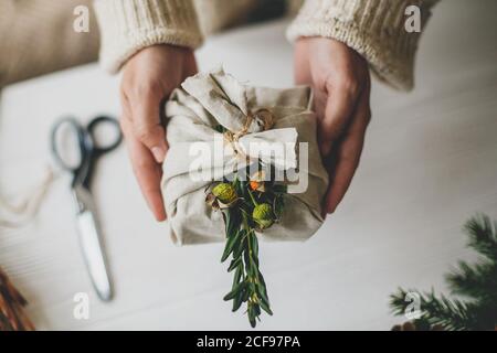 Zero waste Christmas gift. Hands holding stylish present wrapped in linen fabric with natural green branch on rustic background  with pine cones and s Stock Photo