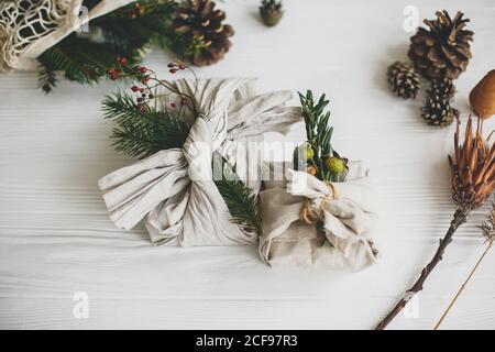 Stylish christmas gifts wrapped in linen fabric, decorated with natural green branch on white rustic table background with pine cones and herbs. Zero Stock Photo