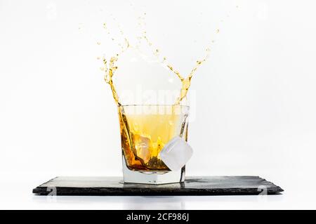 Glass of yellow cold drink with falling ice cube and splashes placed on black slate board on white background Stock Photo