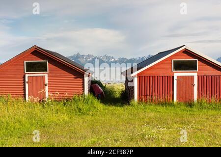 Red wooden buildings located on green meadow on shore with rocky mountains and cloudy sky in background in sunny day in countryside Stock Photo