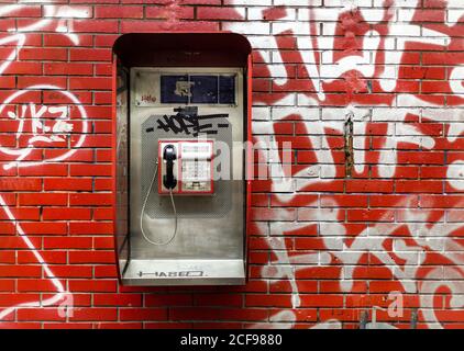 Urban public pay phone hanging on red brick wall with white graffiti on city street Stock Photo