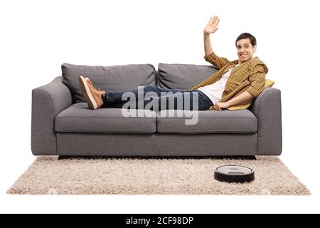 Guy lying on a sofa and waving at the camera while a robot vacuum cleaner is cleaning the carpet isolated on white background Stock Photo