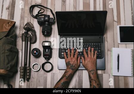 Overhead crop man with tattooed hands typing on laptop keyboard near photography equipment and stationery on lumber surface Stock Photo