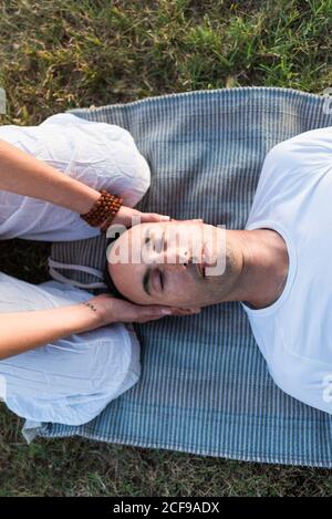 calm couple meditating while man lying on mat with eyes closed and Woman sitting on knees and holding partner by head during looking down in green meadow Stock Photo