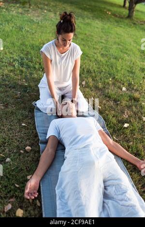 From above of calm couple meditating while man lying on mat with eyes closed and Woman sitting on knees and holding partner by head during looking down in green meadow Stock Photo