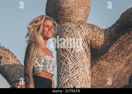 Cheerful young blonde wearing casual summer clothes with tattoos on shoulders resting on wicker seats while looking away on sunny day in summer Stock Photo