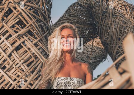 Cheerful young blonde wearing casual summer clothes with tattoos on shoulders resting on wicker seats while looking away on sunny day in summer Stock Photo