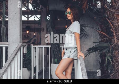 Low angle side view of happy slim female in gray casual shirt and shorts with smartphone touching sunglasses and looking away with interest while leaning on railing at wooden staircase in comfortable resort hotel on Bali Stock Photo